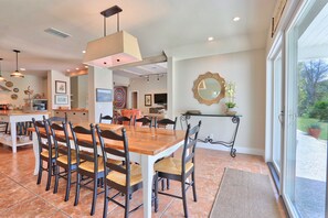 A spacious dining area with a wooden table and chairs, a kitchen in the background, and a large window with a view of the outdoors. - Relaxation awaits you. Open spaces throughout! The Blanco River welcomes you. (Wimberley)