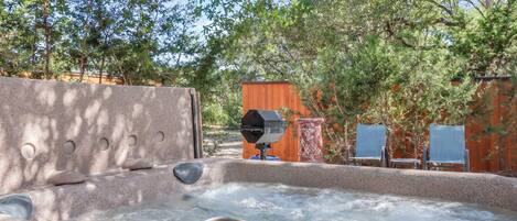 A hot tub with a stone surround, a grill, and two lounge chairs.