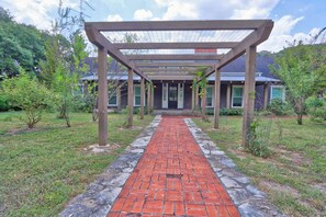 A brick pathway leading to a covered pergola structure with a view of a house in the background. - Relaxation awaits you. Open spaces throughout! The Blanco River welcomes you. (Wimberley)