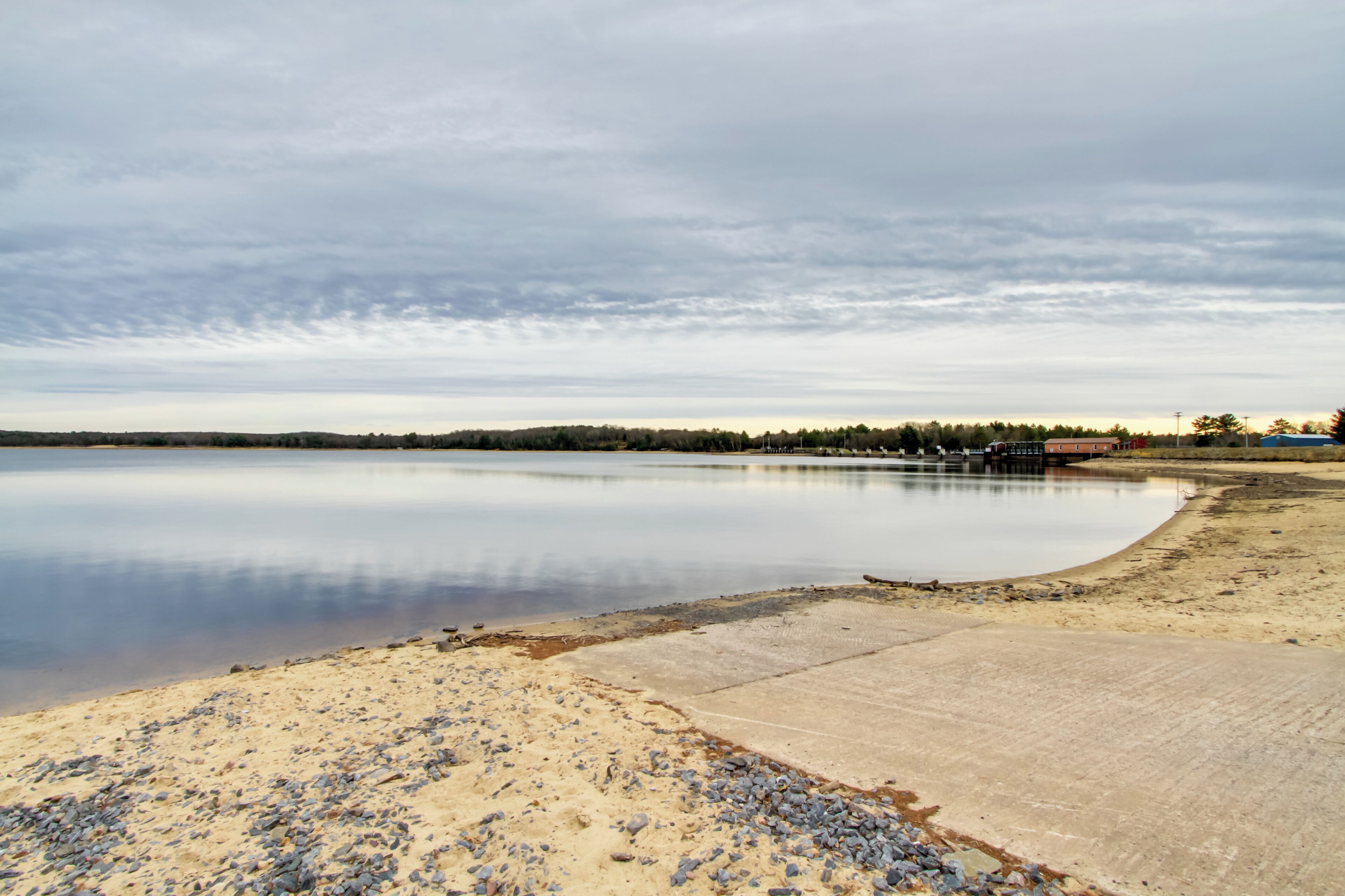 A calm lake with a sandy shore and a concrete path leading to a dock.