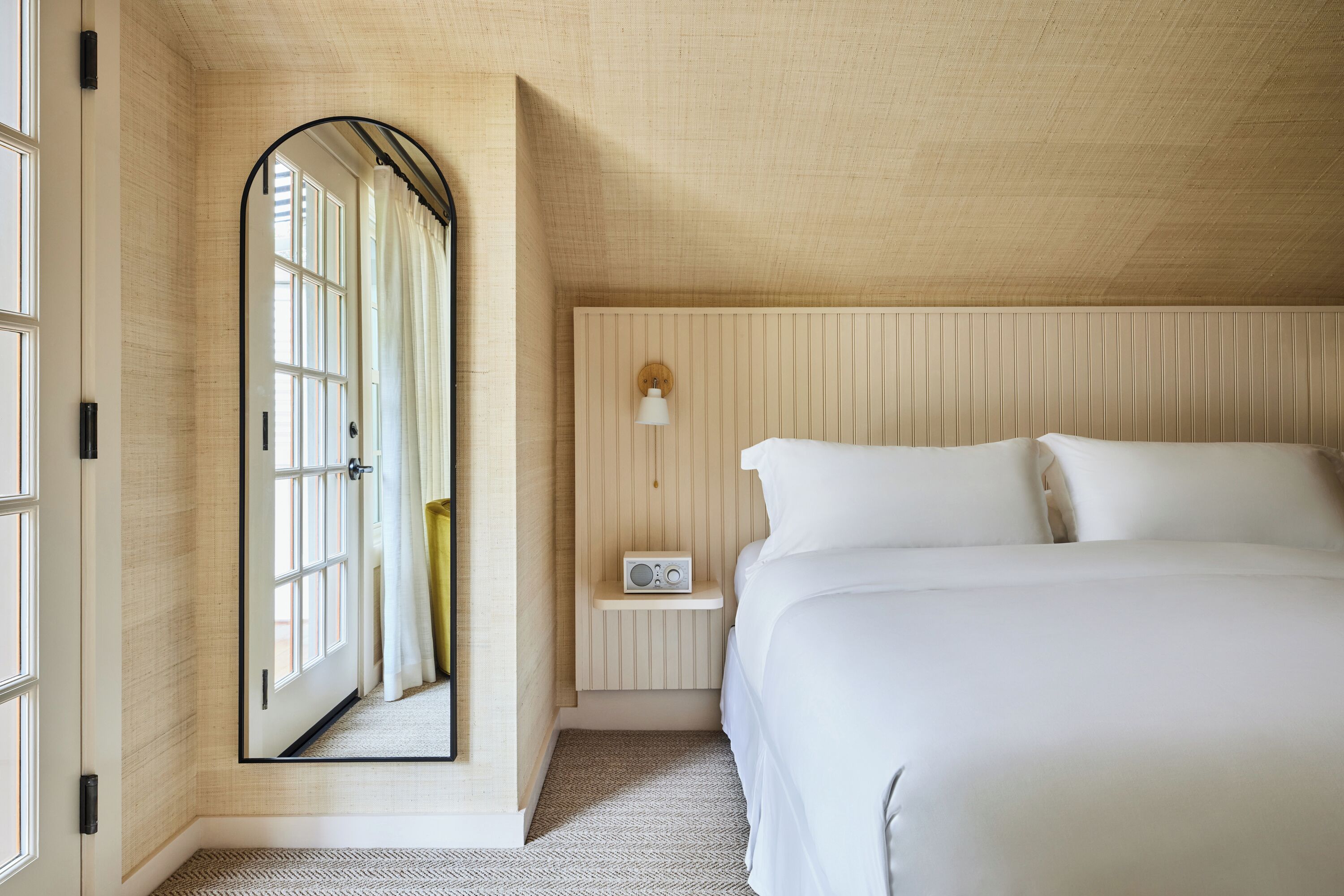 A bedroom with a large mirror, a bedside table with a radio, and a bed with white linens.