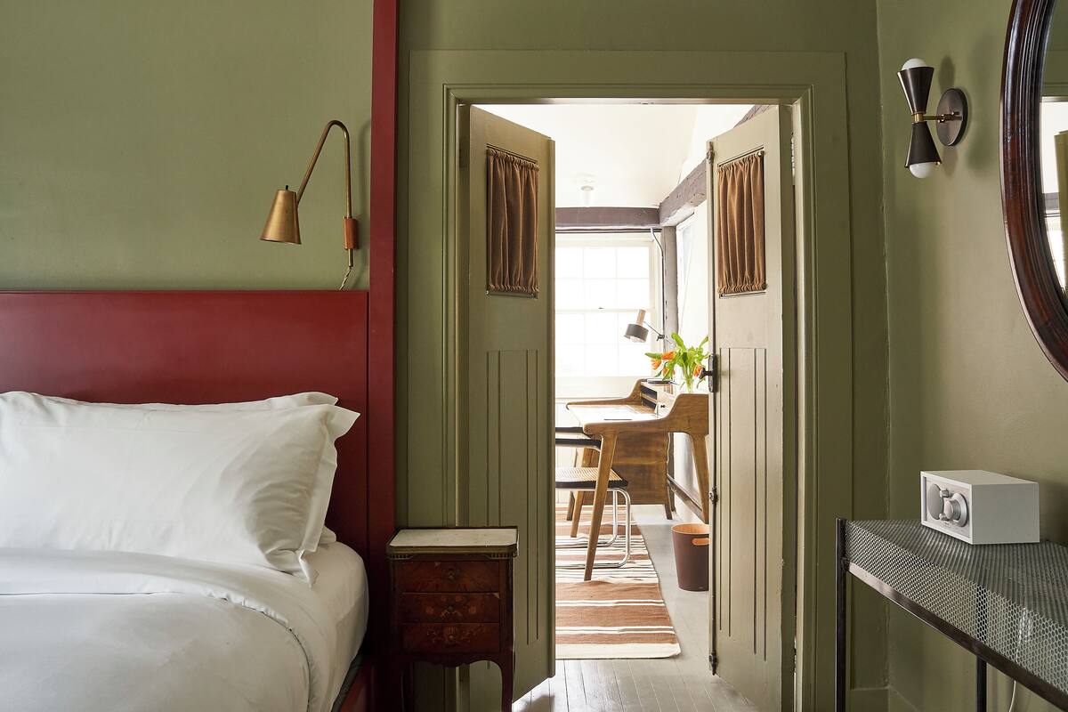 Une chambre à coucher comprenant un lit, une table de chevet, une commode, un miroir et une porte donnant sur un espace cuisine.