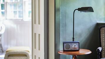 Quarto com piso de madeira, uma pequena mesa redonda com um rádio e uma luminária, uma cadeira estampada e uma cadeira de vime ao fundo.