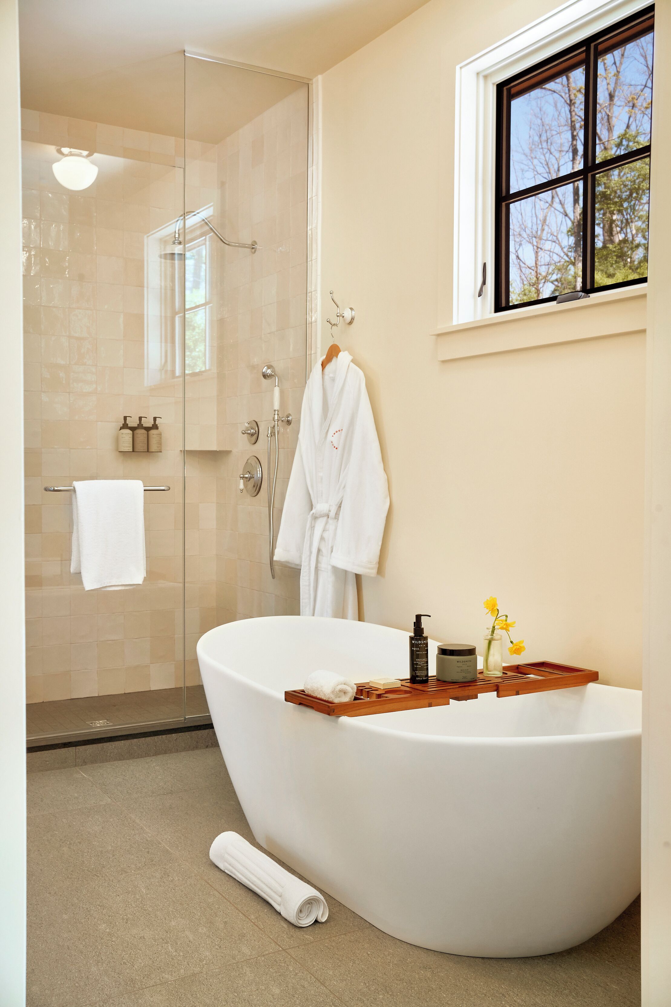 Ein modernes Badezimmer mit freistehender Badewanne, einer Duschkabine aus Glas und einem Fenster mit Blick auf Bäume.