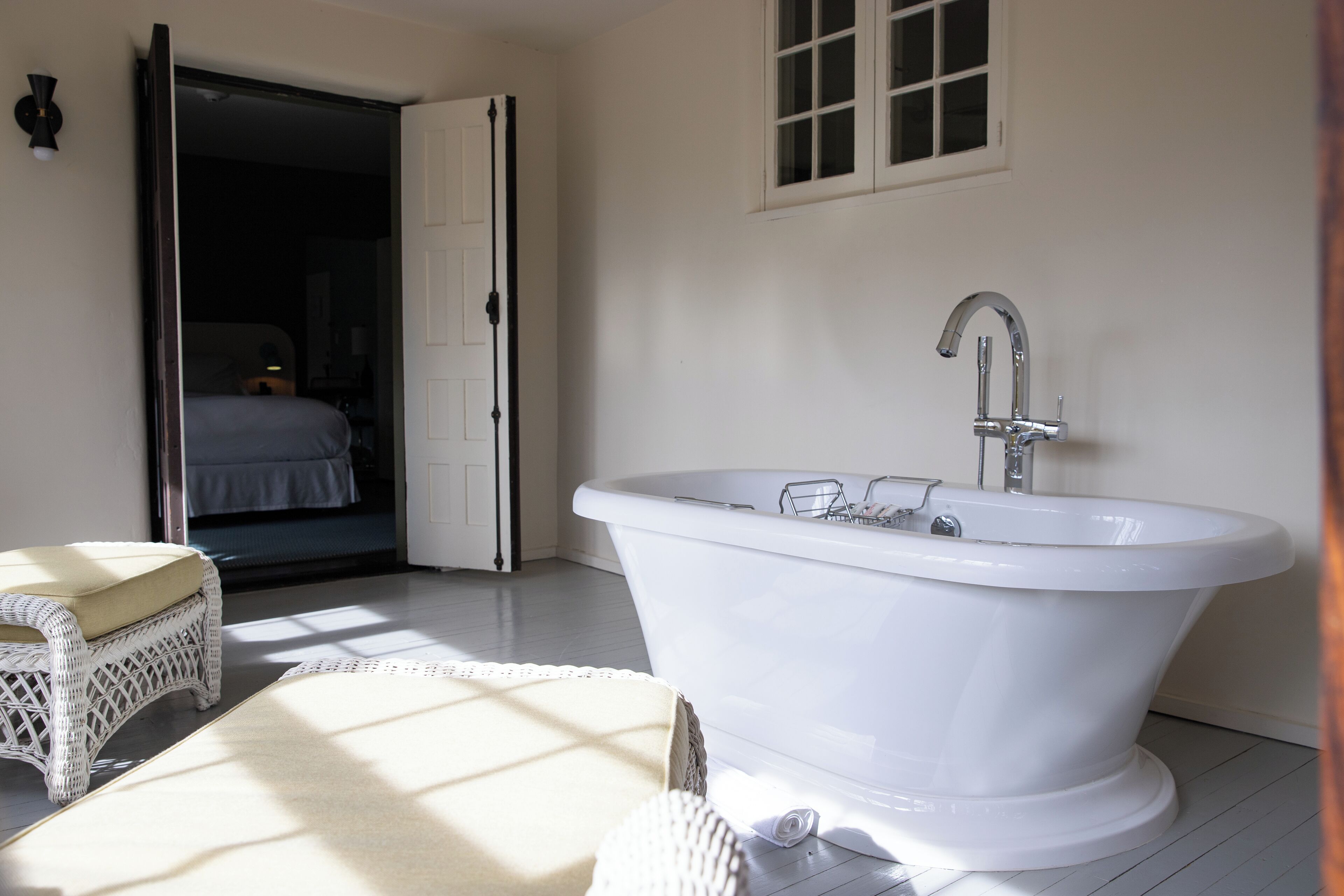 A bathroom with a freestanding bathtub, a wicker chair, and a door leading to another room.