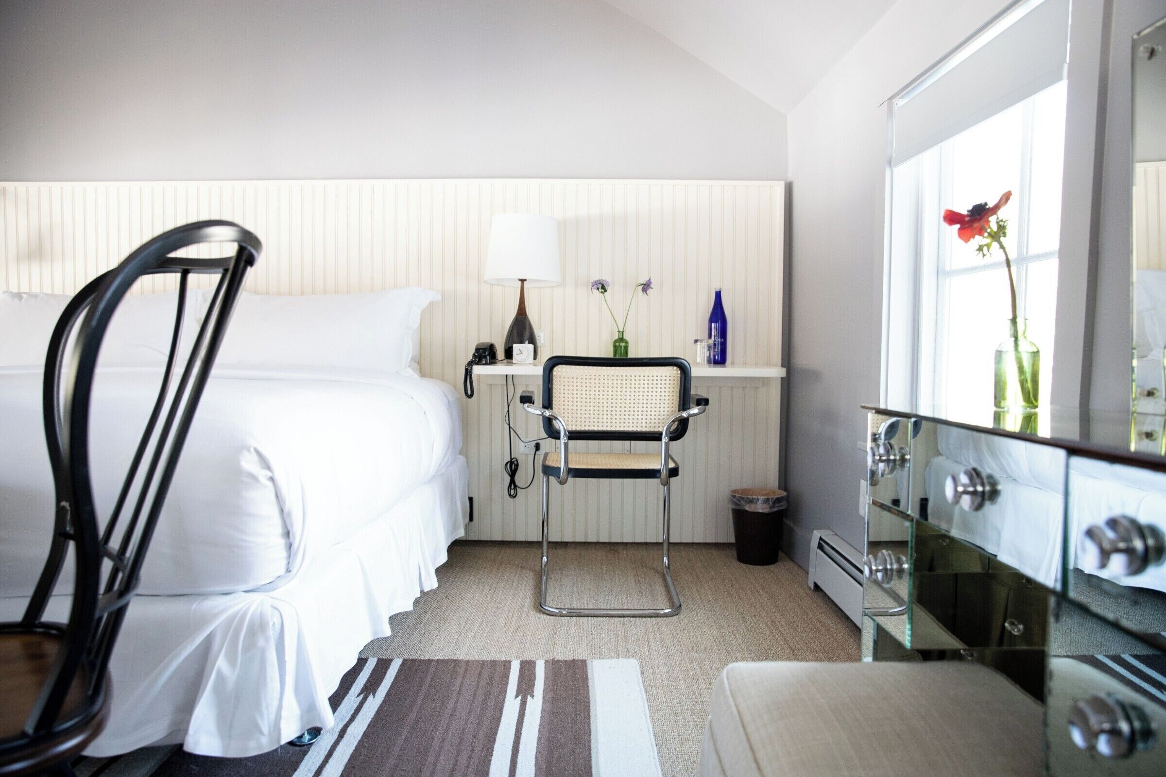 Una habitación de hotel con cama, una silla, un escritorio con lámpara, cómoda y ventana con un jarrón de flores.