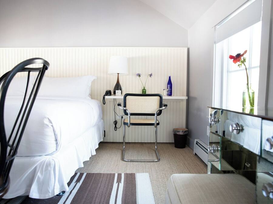 Quarto de hotel com cama, poltrona, escrivaninha com luminária, cômoda e janela com um vaso de flores.