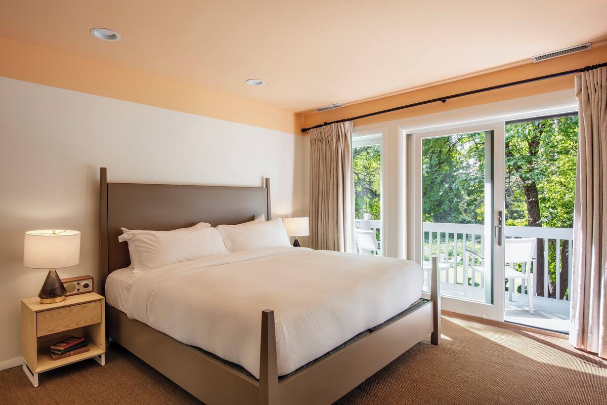 Un lit bien fait, avec du linge de lit blanc, une table de chevet avec une lampe, et une vue sur un balcon doté d’une balustrade blanche et de plantes vertes à l’extérieur.
