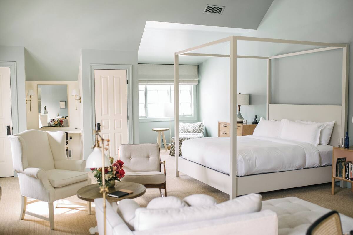 Une chambre à coucher avec un lit à baldaquin, un coin salon avec un fauteuil et une table, et un grand miroir.
