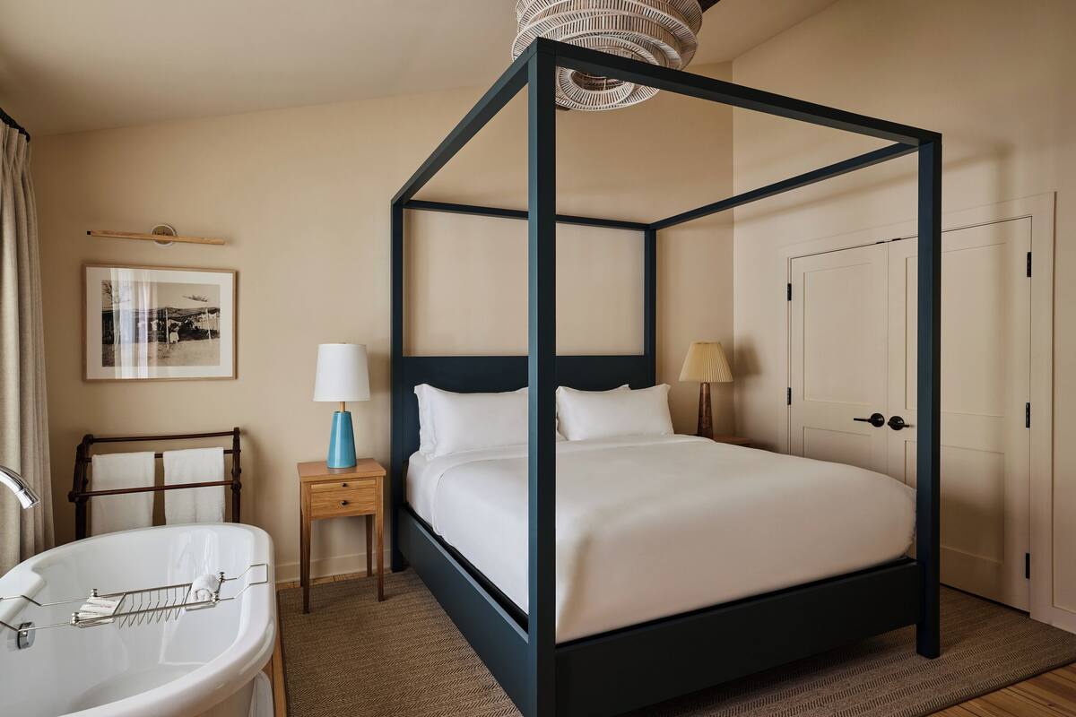 Un lit à baldaquin avec du linge de lit blanc, une table de chevet avec une lampe, un tableau encadré au mur et une salle de bain avec une baignoire et un porte-serviettes.