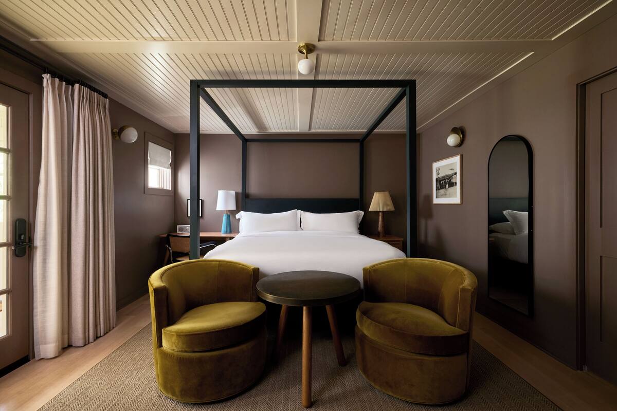 Une chambre d’hôtel avec un lit à baldaquin, deux fauteuils, une petite table ronde et un grand miroir au mur.