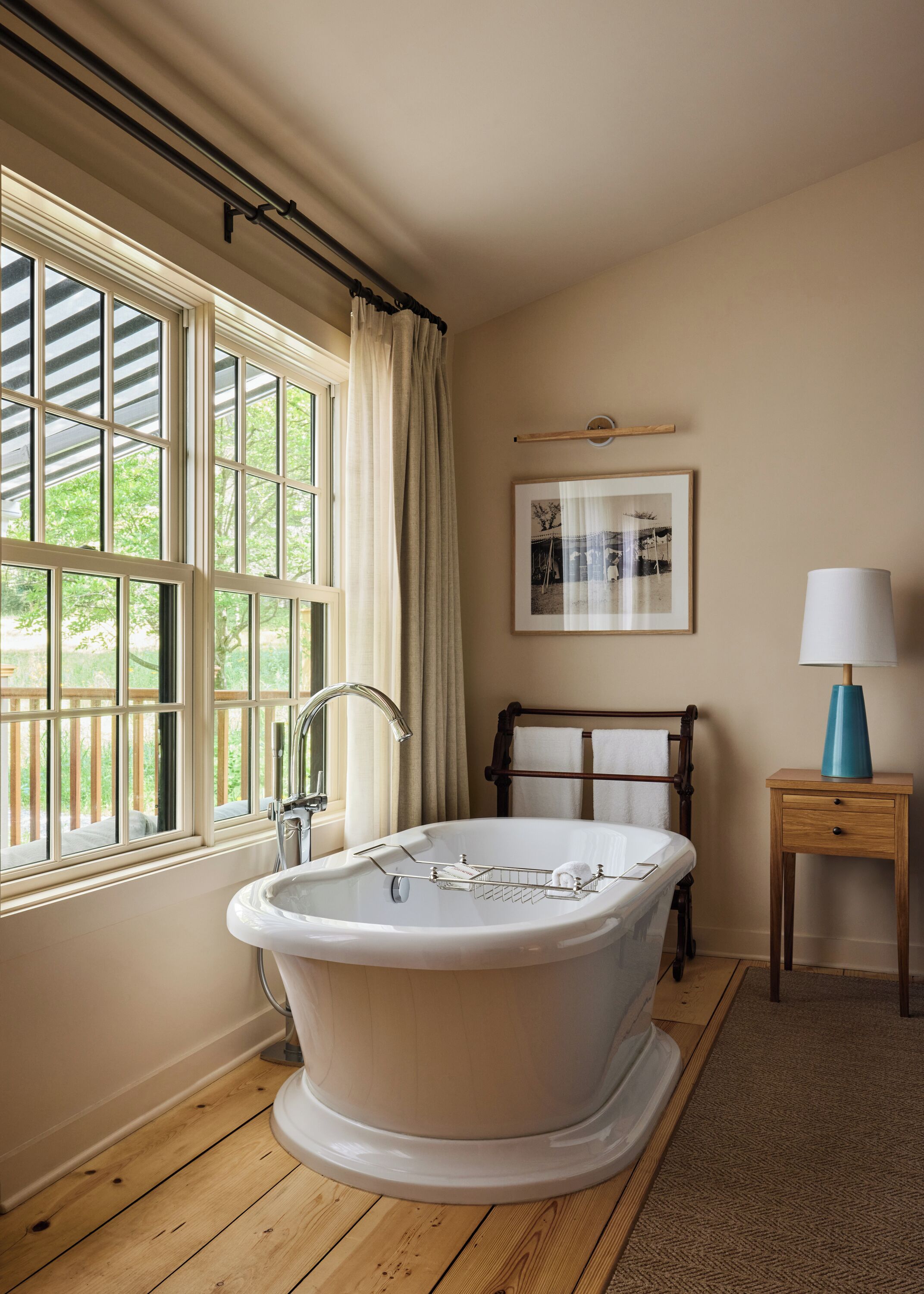 A bathroom with a freestanding bathtub, a wooden floor, a window with curtains, a framed picture on the wall, and a bedside table with a lamp.