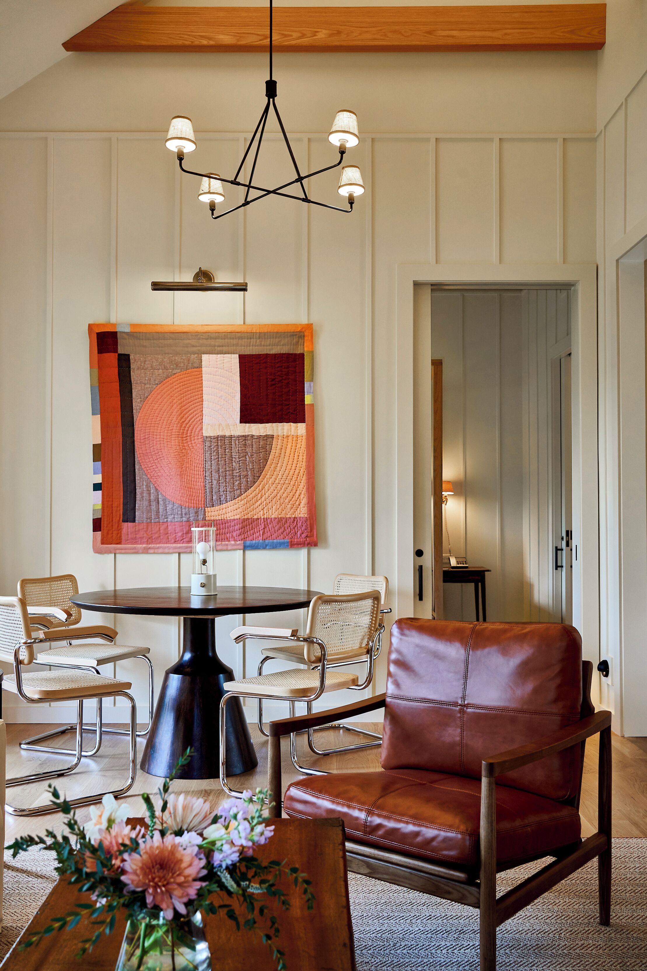 Een eetruimte met een ronde tafel, vier stoelen, een rode lederen fauteuil, een wanddecoratie en een hanglamp.