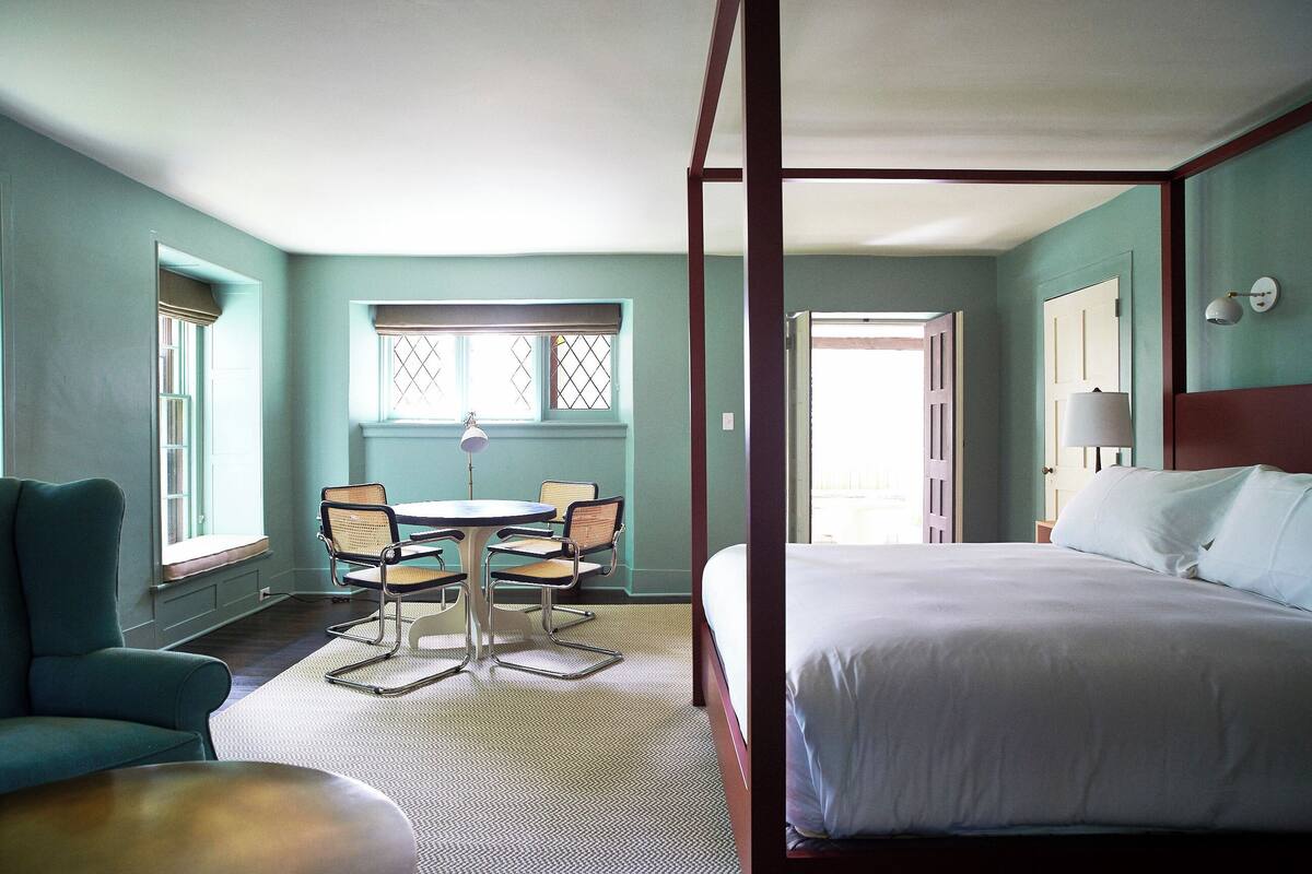 Une chambre d’hôtel avec un lit à baldaquin, une table à manger avec des chaises et une fenêtre donnant sur un beau paysage.