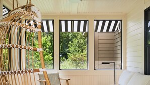A room with a hanging rattan chair, a wooden sofa, a small round table, and a ceiling fan. Large windows provide views of greenery outside. - Troutbeck (Amenia)