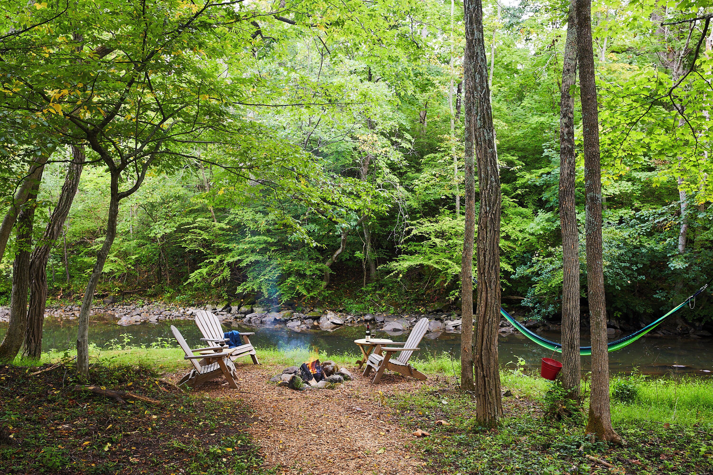 Un camping con dos sillas Adirondack, un pequeño hogar para fogata y una hamaca cerca de una zona boscosa con un arroyo.
