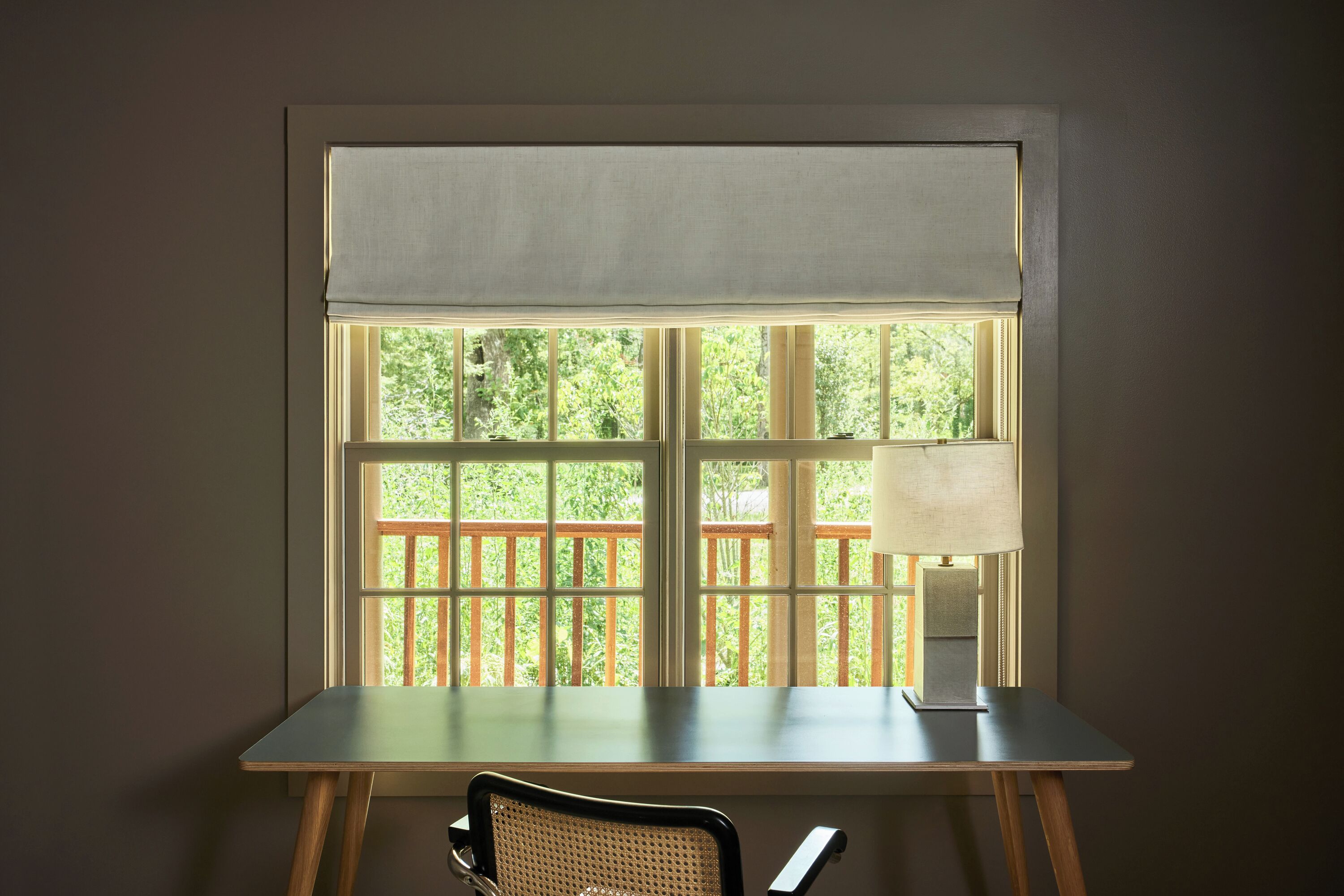 A room with a desk, chair, and window with a view of a wooden deck and greenery.