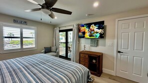 A bedroom with a bed, a television, a ceiling fan, a window with blinds, and a door.