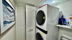 A laundry room with a stacked washer and dryer, a door with horizontal slats, and a shelf with cleaning supplies.