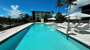 A clear blue pool with lounge chairs and umbrellas, surrounded by palm trees and a building in the background.