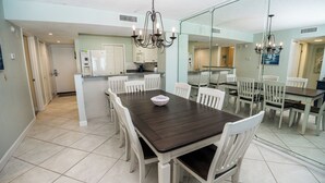 A dining area with a dark wooden table and white chairs, a kitchen in the background, and a chandelier. - E805 Great Gulf View, 3 pools, Gym & Tennis Court (Panama City)