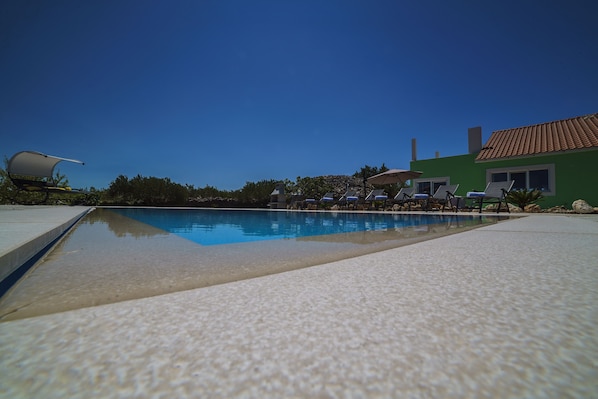 Una piscina con sillas de descanso y sombrillas, rodeada por un edificio verde con tejado de tejas.