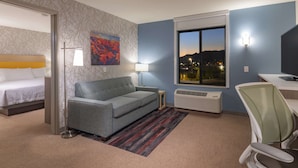 A hotel room with a bed, a gray sofa, a desk with a chair, a window with a view of the city, and a painting on the wall.