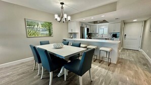 A modern kitchen with white cabinets, a central island, and a dining area with a white table and blue chairs. - Sandpiper Beach #202 ~ Reimagined. Rebuilt. Better Than Ever (Sanibel)
