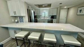 A modern kitchen with a large island, bar stools, stainless steel appliances, and white cabinetry. - Sandpiper Beach #202 ~ Reimagined. Rebuilt. Better Than Ever (Sanibel)