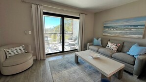 A living room with a sofa, a coffee table, a chair, and a view of a balcony with outdoor furniture. - Sandpiper Beach #202 ~ Reimagined. Rebuilt. Better Than Ever (Sanibel)