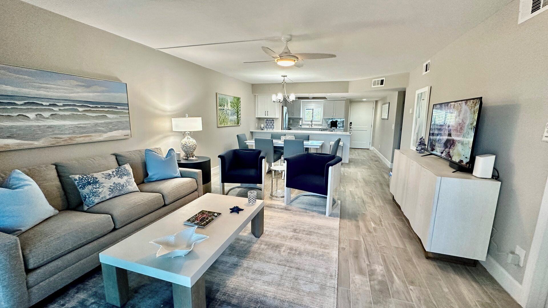 A modern living room with a sofa, coffee table, and chairs. There is a dining area with a table and chairs, and a kitchen in the background.