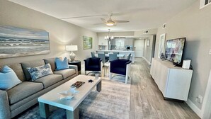 A modern living room with a sofa, coffee table, and chairs. There is a dining area with a table and chairs, and a kitchen in the background. - Sandpiper Beach #202 ~ Reimagined. Rebuilt. Better Than Ever (Sanibel)