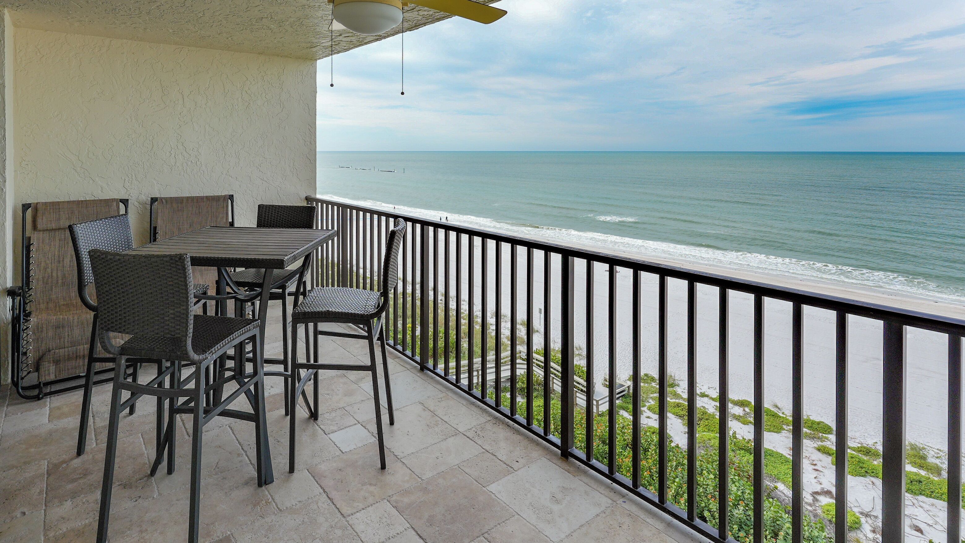 A balcony with a table and chairs, overlooking a beach with the ocean and a clear sky.