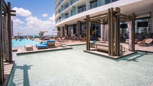 A pool area with lounge chairs and umbrellas, a large building with balconies, and a clear sky.