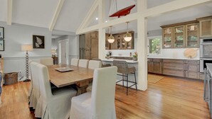 Uma ampla área de jantar com uma mesa de madeira e cadeiras estofadas brancas, uma cozinha com armários de madeira e iluminação pendente, e um alto teto com vigas aparentes.