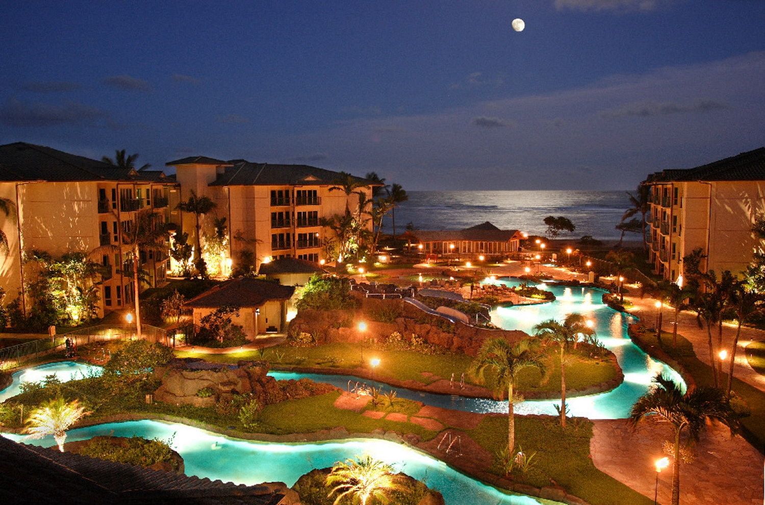A resort with multiple swimming pools, a large building, and a view of the ocean at night.
