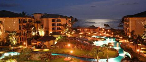 A resort with multiple swimming pools, a large building, and a view of the ocean at night.