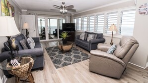 A spacious living room with a grey sofa, a beige armchair, and a television on a stand. There is a large window with blinds and a ceiling fan. - D601 Direct Gulf View, 3 pools, Tennis Court (Panama City)