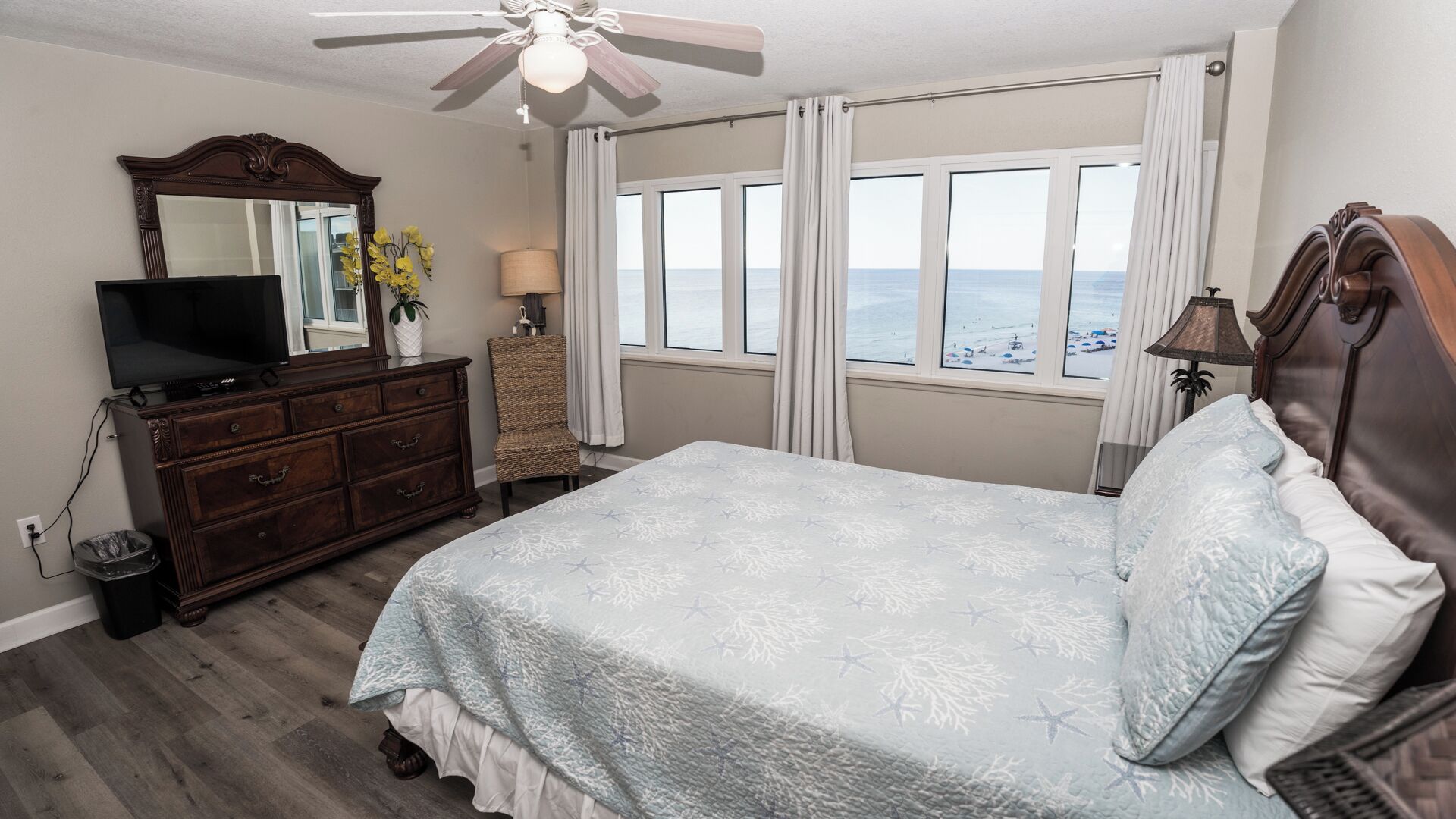 A bedroom with a bed, a dresser, a television, a ceiling fan, and a view of the beach through large windows.