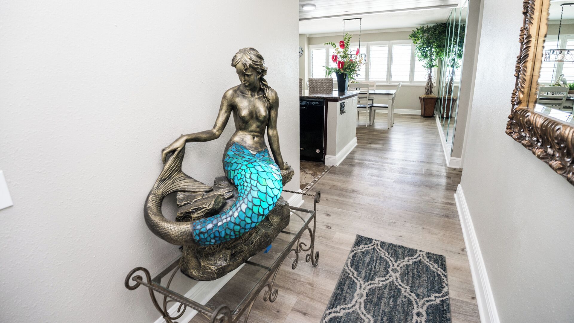 A hallway with a decorative mermaid sculpture on a stand, a kitchen area with a dining table and chairs, and a large mirror on the right.