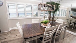 A dining area with a wooden table, white chairs, and a view of the ocean through large windows. - D601 Direct Gulf View, 3 pools, Tennis Court (Panama City)