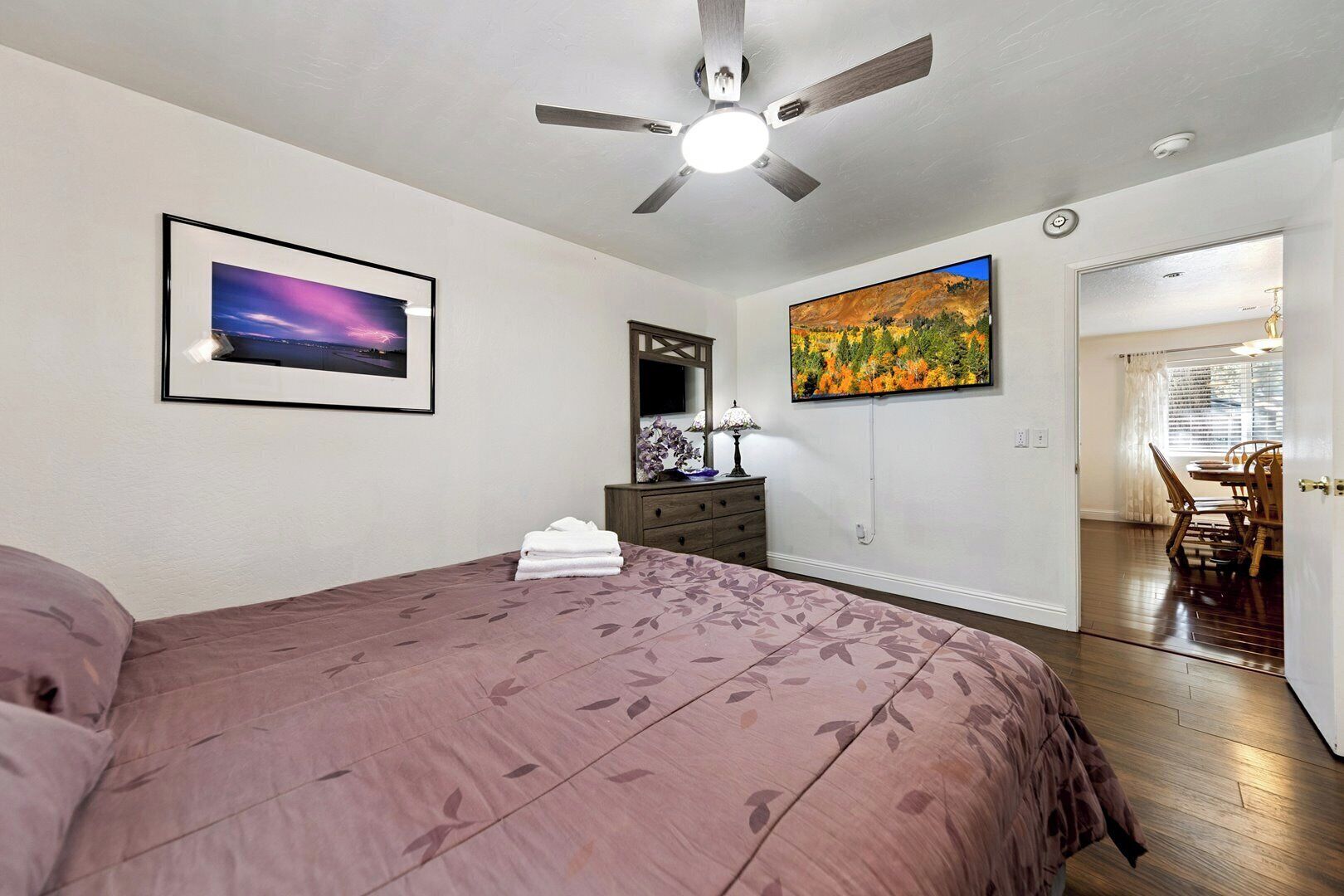 Quarto com cama, ventilador de teto, mesa de cabeceira, luminária, pintura e porta que dá para uma sala de jantar.