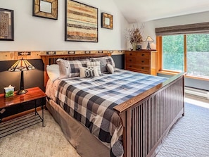 A bedroom with a large bed, a nightstand, a dresser, and a window with a view of greenery.