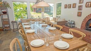 Zona pranzo con un lungo tavolo in legno apparecchiato per sei persone, circondato da sedie di legno. È presente un camino in mattoni e un'ampia finestra che offre vista sul verde circostante.