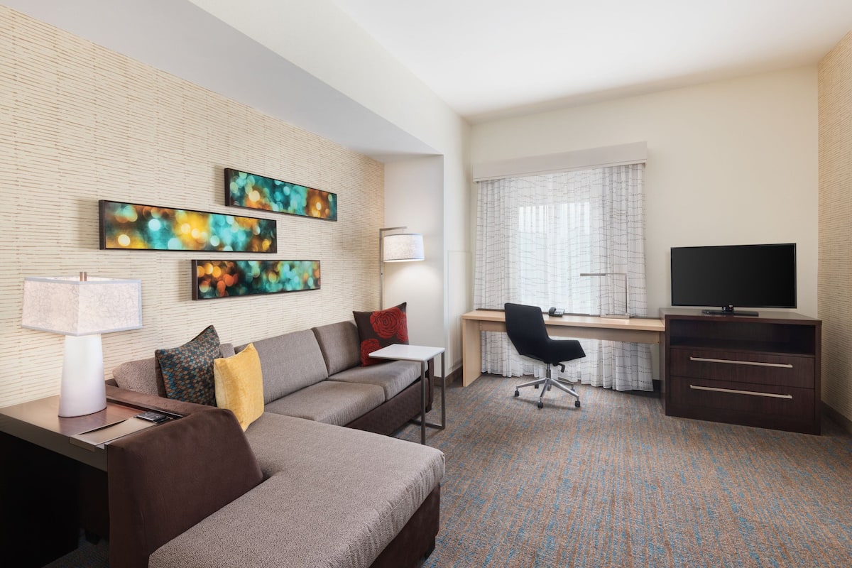 A modern hotel room with a sofa, desk, TV, and artwork on the wall.