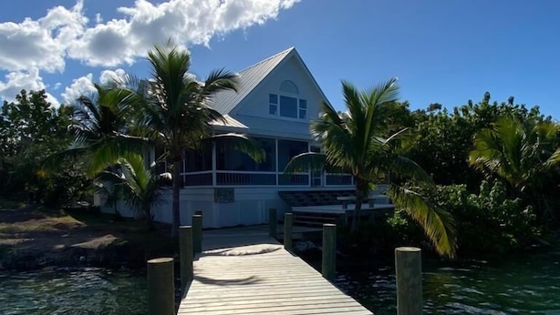A wooden dock leading to a house with a white roof, surrounded by palm trees and clear blue water. - Hope Town harbor period home with lighthouse view (Hope Town)