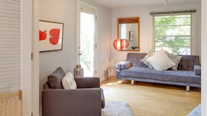 Una sala moderna con un sofá gris, un sillón más pequeño y una alfombra blanca. Hay un ventanal con vistas al exterior, lleno de vegetación.