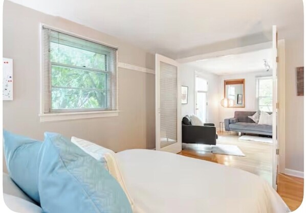 A bedroom with a bed, a window, and a view of a living area with a sofa and a chair.