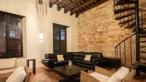 Una sala de estar acogedora con un sofá de cuero negro, sillas de mimbre y una mesita de centro. Se aprecia una pared de ladrillo y vigas de madera al fondo.