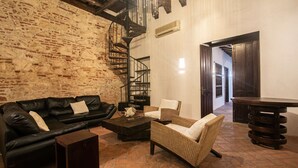 Una sala de estar acogedora con pared de ladrillo, un sofá de cuero negro, un sillón de mimbre, una mesa de centro de madera y una escalera en espiral.